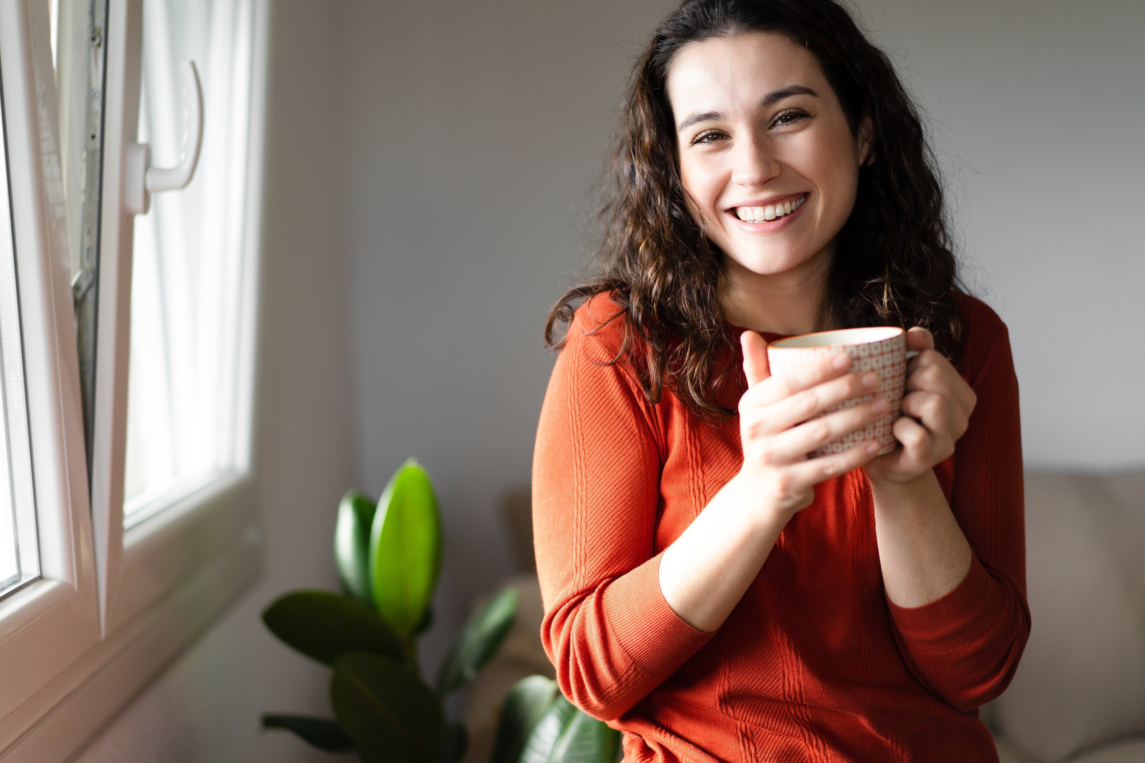 Woman in orange top smiling and holding a cup with a green plant behind her. 